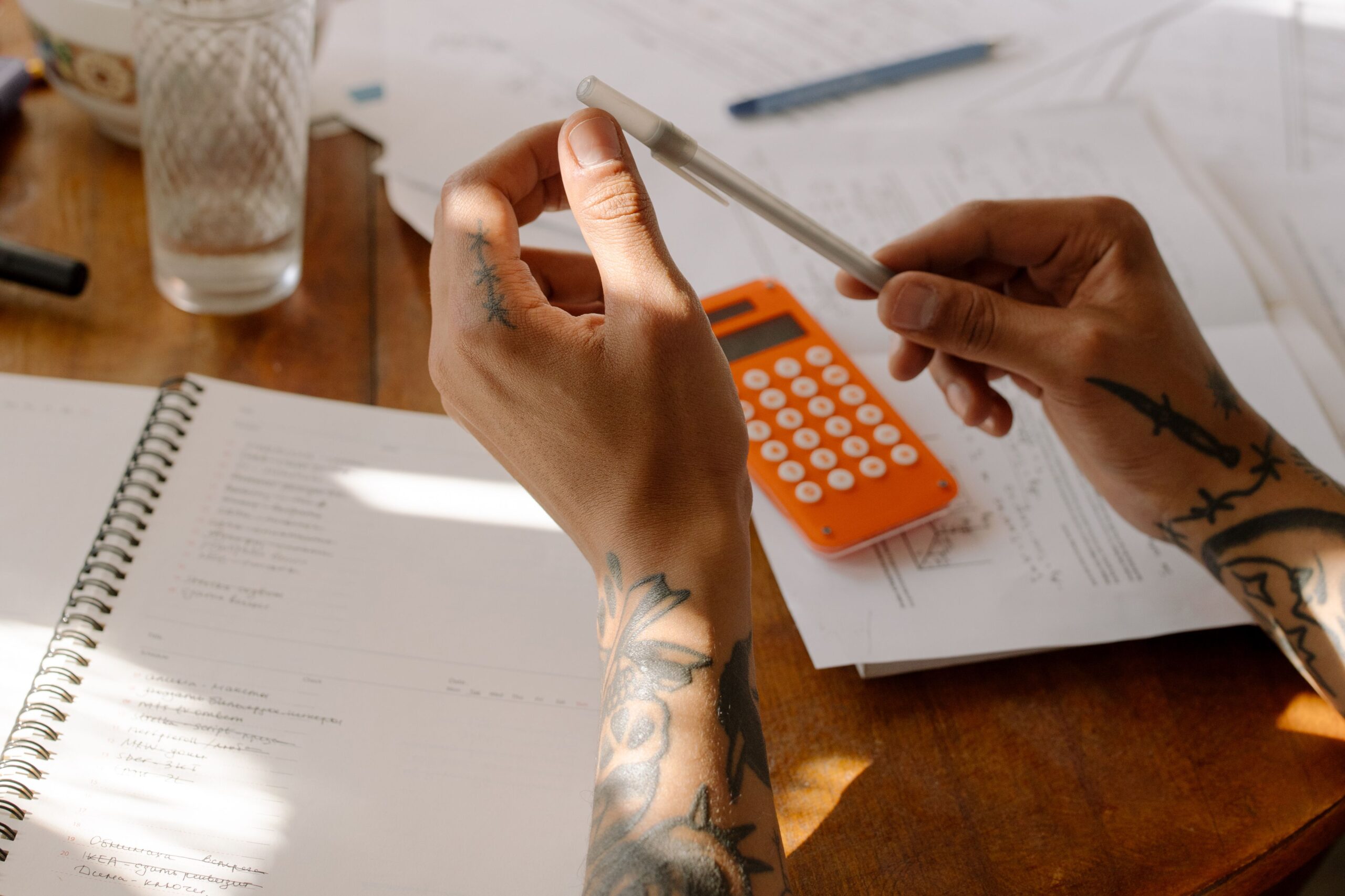 Close-up of tattooed hands holding a pen next to a calculator and notebooks.